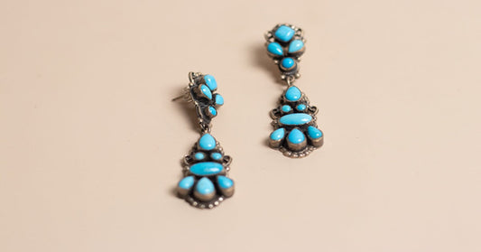 A pair of silver drop earrings with bright blue stones resting on a plain beige surface, with soft shadows around them.