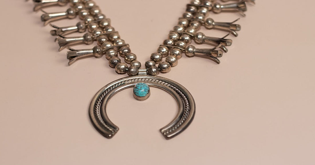 A silver beaded necklace with a horseshoe-shaped pendant and a small turquoise stone on a beige surface.