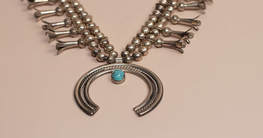 A silver beaded necklace with a horseshoe-shaped pendant and a small turquoise stone on a beige surface.