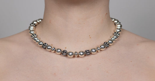 A person wearing a silver beaded necklace against a plain gray background, with the neck and shoulders visible.