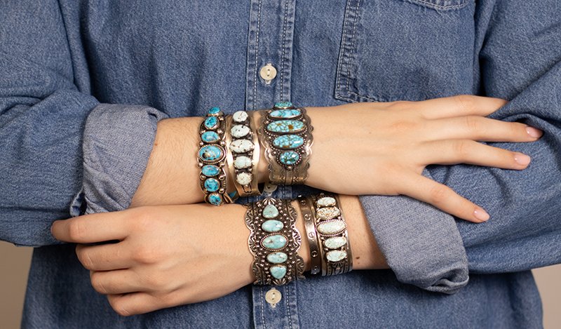 A person's crossed arms wearing stacked silver and turquoise bracelets over a blue denim button-up shirt.