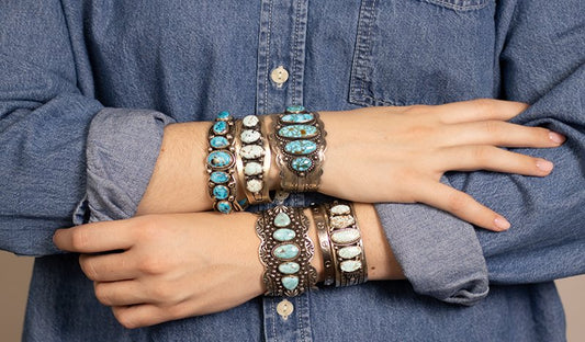 A person's crossed arms wearing stacked silver and turquoise bracelets over a blue denim button-up shirt.